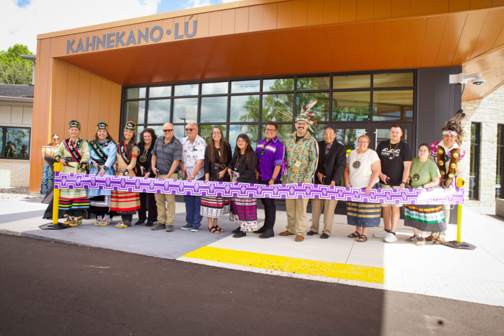 Kahnekano·lú staff members and Oneida Royalty standing in front of the Kahnekanó·lú Cultural Center during a ribbon-cutting ceremony, holding a purple patterned ribbon outside the building entrance.